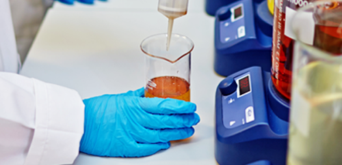 a hand in a glove holding a chemical vessel and filling it with an orange e-liquid, scales standing near the vessel
