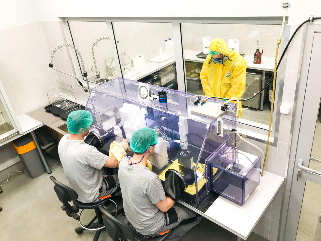 3 people at nicotine workshop, 2 at the desk, 1 behind the glass, wearing a safety uniform, mask, gloves and googles