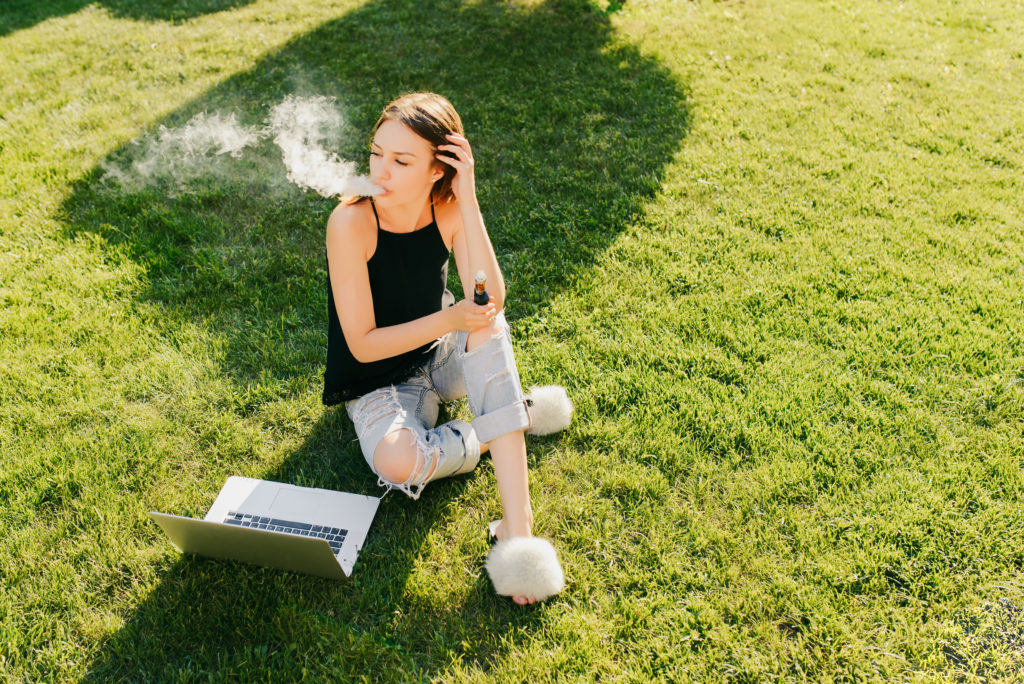 a woman vaping, sitting on grass with a laptop